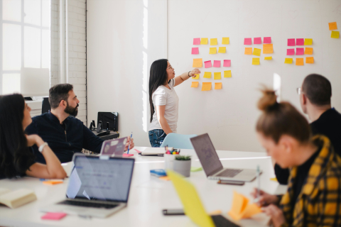 Woman placing sticky notes on wall with others seated around table.